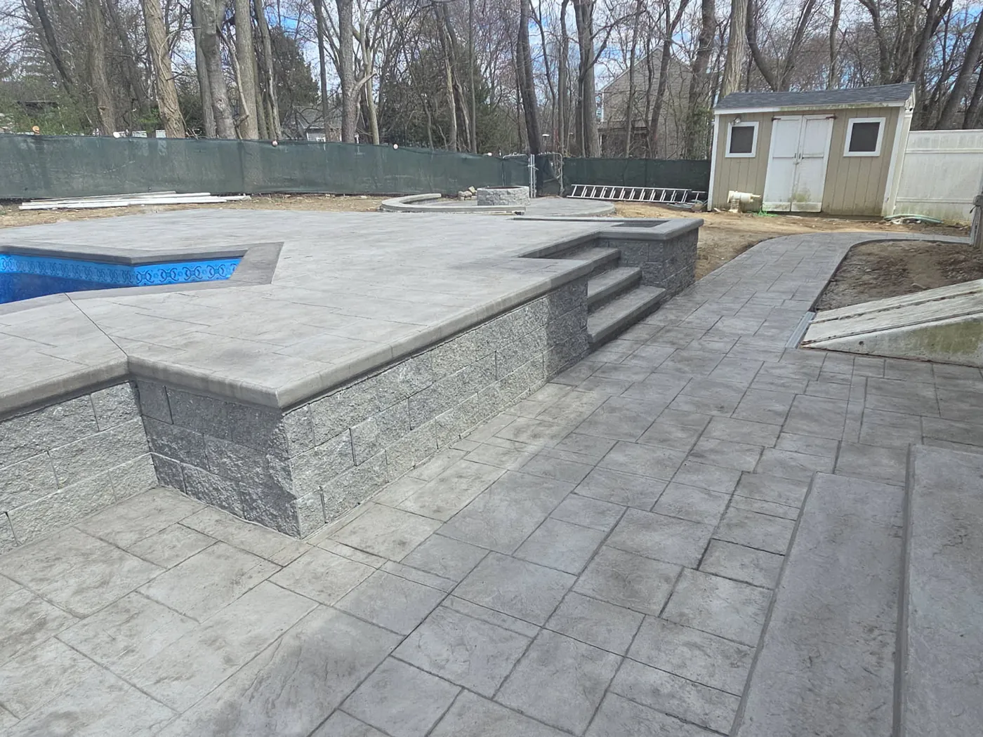 Stamped concrete patio and block steps beside a pool with a garden shed behind