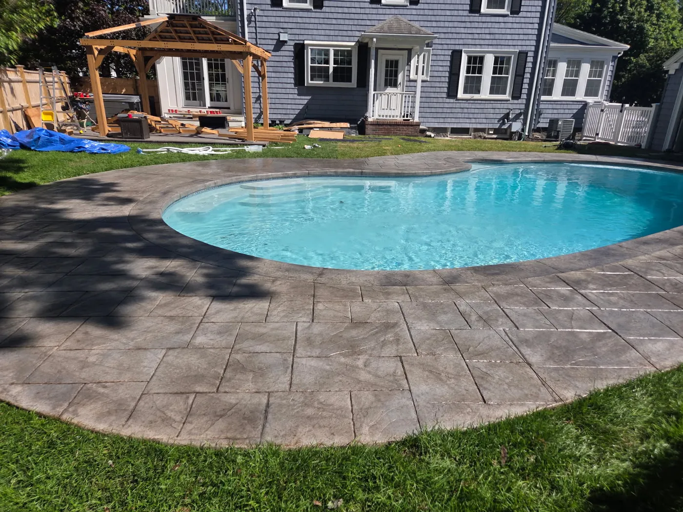 Stamped concrete pool surround beside a wooden gazebo and a blue sided house